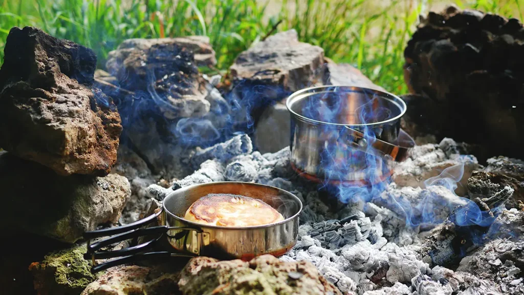 En stekpanna med en pannkaka i och en kastrull som står på en glödbäd i en eldstad ute i naturen.